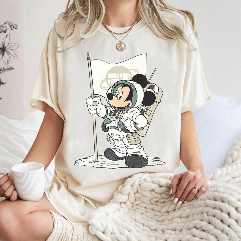 May include: A cream colored t-shirt with a cartoon image of Mickey Mouse dressed as an astronaut standing on the moon. Mickey is holding a flag with a circle and a planet on it.