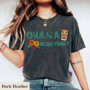 May include: Dark heather gray t-shirt with the text "OHANA MEANS FAMILY" in teal and orange. The shirt features a floral design and a tiki graphic, evoking a tropical theme.