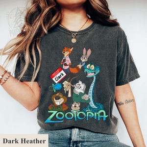 Puede incluir: Camiseta gris oscuro con un dise&ntilde;o gr&aacute;fico colorido de la pel&iacute;cula Zootopia. El dise&ntilde;o presenta personajes como un zorro, un conejo y una serpiente. La palabra "Zootopia" est&aacute; escrita en azul y verde.