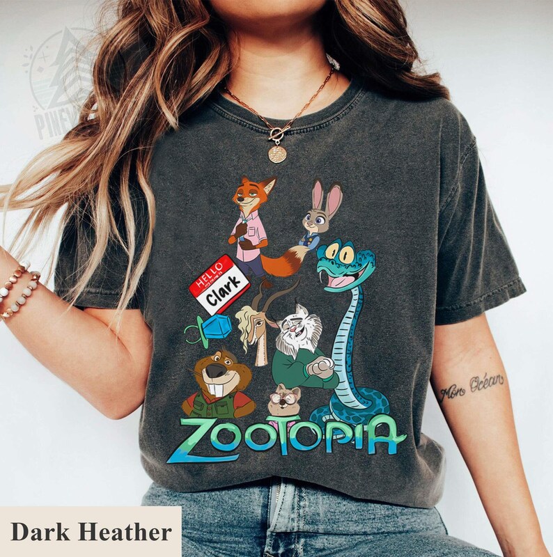 Puede incluir: Camiseta gris oscuro con un dise&ntilde;o gr&aacute;fico colorido de la pel&iacute;cula Zootopia. El dise&ntilde;o presenta personajes como un zorro, un conejo y una serpiente. La palabra "Zootopia" est&aacute; escrita en azul y verde.