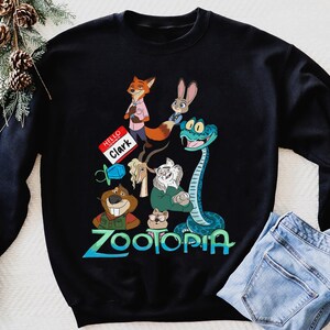 Puede incluir: Sudadera negra con un dise&ntilde;o colorido con personajes de la pel&iacute;cula animada Zootopia. El dise&ntilde;o incluye un zorro, un conejo y una serpiente. La palabra "Zootopia" est&aacute; escrita en azul y verde. Una etiqueta con el nombre dice "Hello Clark".