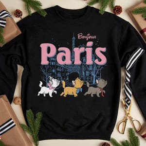 Puede incluir: Sudadera negra con la palabra "Par&iacute;s" en rosa y "Bonjour" encima. El dise&ntilde;o incluye la Torre Eiffel y tres gatitos de dibujos animados. La sudadera est&aacute; rodeada de elementos festivos, como ramas de pino y regalos envueltos.