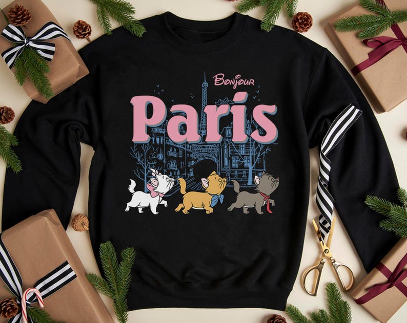 Puede incluir: Sudadera negra con la palabra "Par&iacute;s" en rosa y "Bonjour" encima. El dise&ntilde;o incluye la Torre Eiffel y tres gatitos de dibujos animados. La sudadera est&aacute; rodeada de elementos festivos, como ramas de pino y regalos envueltos.