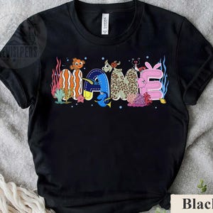 May include: Black t-shirt with the word "GAME" in colorful, cartoon-style lettering. The letters are designed with aquatic themes, including fish and coral, creating a playful and vibrant design. The shirt is made of a soft material.