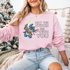 May include: A light pink sweatshirt featuring a cartoon Stitch character holding a green lightsaber. The text "MAY THE STITCH BE WITH YOU" is printed in black. The sweatshirt is a long-sleeved crew neck.