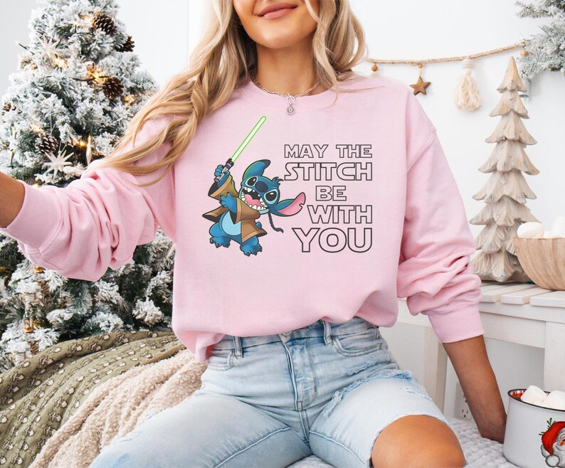 May include: A light pink sweatshirt featuring a cartoon Stitch character holding a green lightsaber. The text "MAY THE STITCH BE WITH YOU" is printed in black. The sweatshirt is a long-sleeved crew neck.