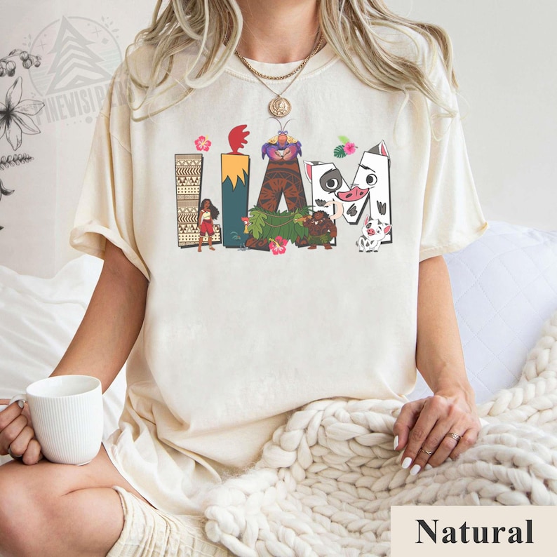 May include: Cream-coloured t-shirt featuring the word "LAVA" in colourful, cartoon-style lettering. The letters are decorated with images of characters and elements from a popular animated movie. The shirt is made of a soft, comfortable material.
