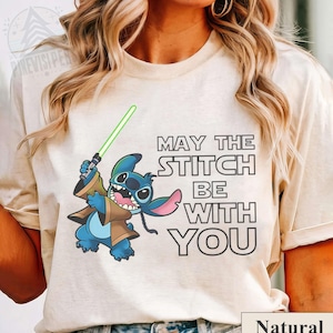 May include: A natural-colored t-shirt featuring a cartoon Stitch character holding a green lightsaber. The text "MAY THE STITCH BE WITH YOU" is printed on the shirt. The word "Natural" is printed at the bottom.
