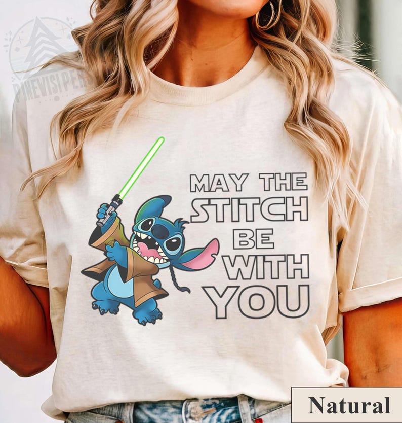 May include: A natural-colored t-shirt featuring a cartoon Stitch character holding a green lightsaber. The text "MAY THE STITCH BE WITH YOU" is printed on the shirt. The word "Natural" is printed at the bottom.