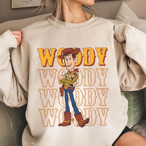 Puede incluir: Sudadera beige con un dise&ntilde;o del personaje Woody de Toy Story. La palabra "WOODY" est&aacute; impresa en un patr&oacute;n repetido en amarillo y naranja. Woody aparece con su atuendo de vaquero, con sombrero, botas y pistolera.
