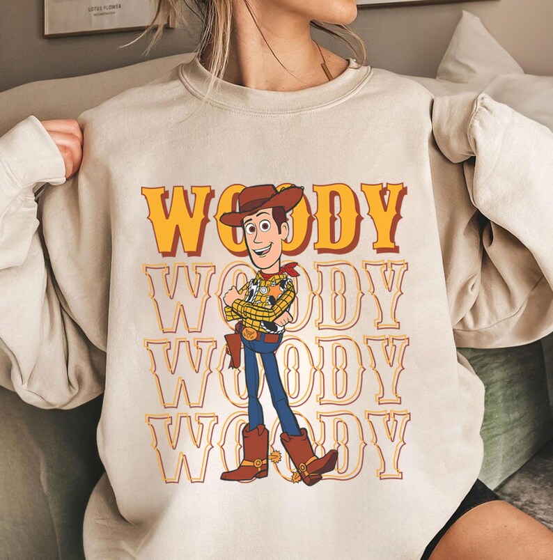 Puede incluir: Sudadera beige con un dise&ntilde;o del personaje Woody de Toy Story. La palabra "WOODY" est&aacute; impresa en un patr&oacute;n repetido en amarillo y naranja. Woody aparece con su atuendo de vaquero, con sombrero, botas y pistolera.