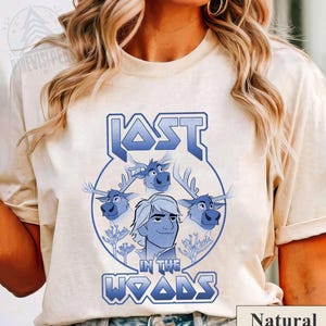 May include: A light beige t-shirt with a blue graphic design featuring a cartoon character with three reindeer. The text "LOST IN THE WOODS" is in a blue, metallic font.