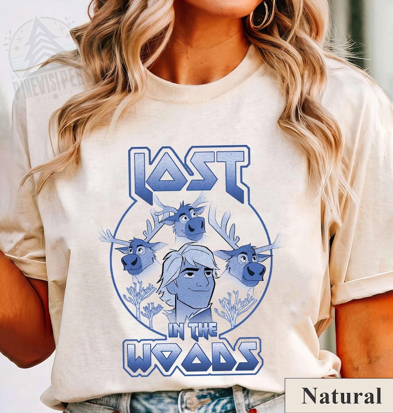 May include: A light beige t-shirt with a blue graphic design featuring a cartoon character with three reindeer. The text "LOST IN THE WOODS" is in a blue, metallic font.
