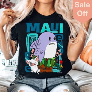 Puede incluir: Una camiseta negra con un gr&aacute;fico de Maui de la pel&iacute;cula Moana. El gr&aacute;fico presenta a Maui como un tibur&oacute;n con un cerdo y un gallo. El texto "MAUI" est&aacute; impreso en azul y verde en la camiseta.