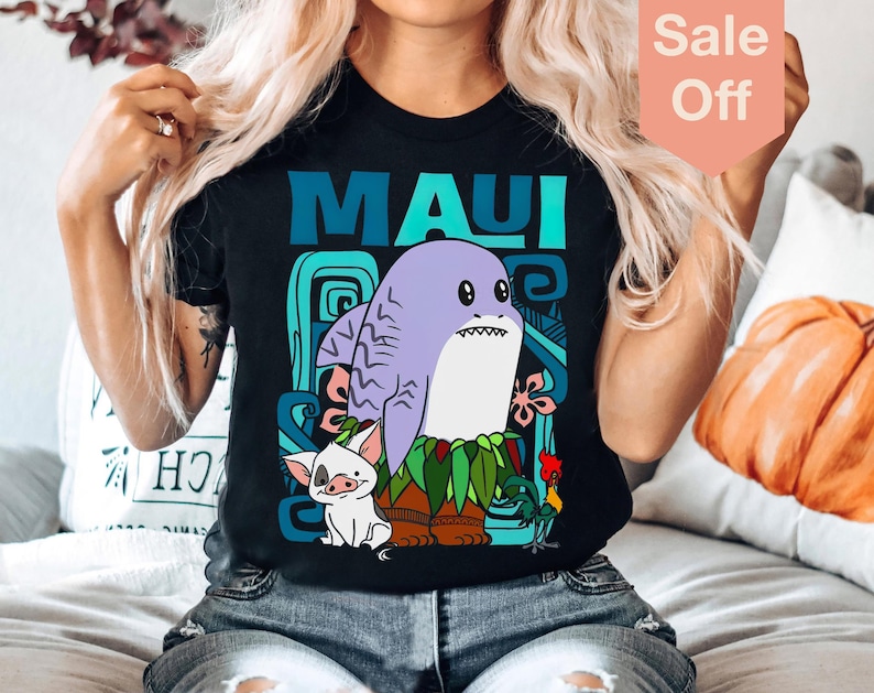 Puede incluir: Una camiseta negra con un gr&aacute;fico de Maui de la pel&iacute;cula Moana. El gr&aacute;fico presenta a Maui como un tibur&oacute;n con un cerdo y un gallo. El texto "MAUI" est&aacute; impreso en azul y verde en la camiseta.