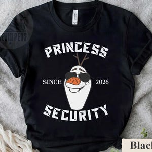 May include: Black t-shirt with a white graphic featuring a cartoon snowman wearing sunglasses and a carrot nose. The text reads "PRINCESS SECURITY SINCE 2026". The shirt is made of a soft material.