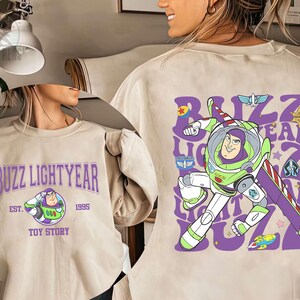 May include: Beige sweatshirt featuring Buzz Lightyear from Toy Story. The front displays "Buzz Lightyear" in purple text with an image of Buzz. The back has a large graphic of Buzz in his spacesuit with the words "Buzz Lightyear" in a repeating pattern.