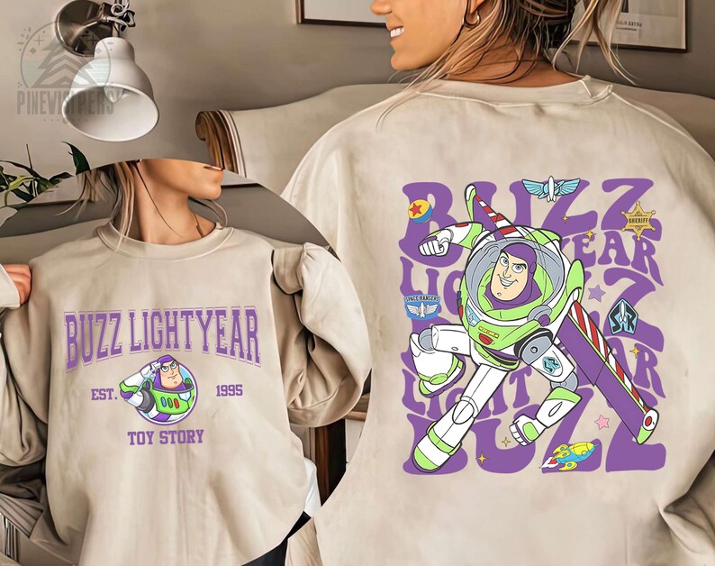 May include: Beige sweatshirt featuring Buzz Lightyear from Toy Story. The front displays "Buzz Lightyear" in purple text with an image of Buzz. The back has a large graphic of Buzz in his spacesuit with the words "Buzz Lightyear" in a repeating pattern.
