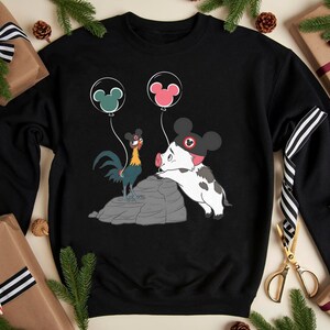 May include: Black sweatshirt with a cartoon rooster and pig design, holding Mickey Mouse ear balloons. The rooster is blue, yellow, and red, and the pig is white and black. Balloons are green and pink.