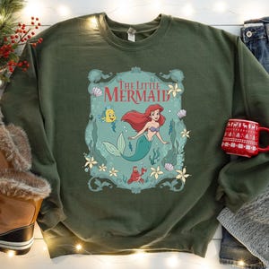 May include: An olive green sweatshirt featuring a graphic of Ariel from The Little Mermaid. The design includes Ariel, Flounder, Sebastian, and the words "The Little Mermaid". The sweatshirt is paired with a red mug and winter boots.
