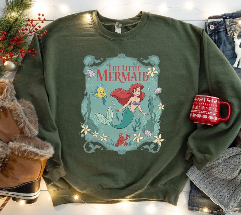 May include: An olive green sweatshirt featuring a graphic of Ariel from The Little Mermaid. The design includes Ariel, Flounder, Sebastian, and the words "The Little Mermaid". The sweatshirt is paired with a red mug and winter boots.