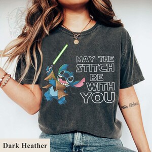 May include: Dark heather gray t-shirt featuring a cartoon Stitch character holding a green lightsaber, with the text "MAY THE STITCH BE WITH YOU" in white. The shirt is a comfortable, casual wear item.