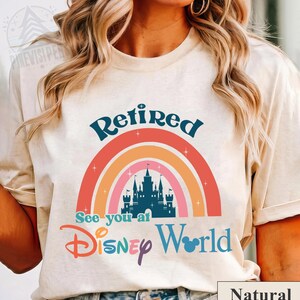 K&ouml;nnte beinhalten: Ein wei&szlig;es T-Shirt mit einem Regenbogen-Design und dem Text "Retired See you at Disney World". Der Regenbogen hat eine Silhouette von Schloss Aschenputtel in der Mitte. Der Text ist in blauer, lila und rosa Schrift. Das Shirt ist mit "Natur" gekennzeichnet.