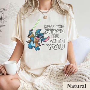 May include: Cream-colored t-shirt featuring a cartoon Stitch character holding a green lightsaber, with the text "MAY THE STITCH BE WITH YOU" in black. The shirt is a natural color.