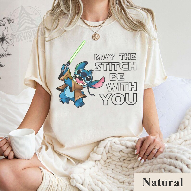 May include: Cream-colored t-shirt featuring a cartoon Stitch character holding a green lightsaber, with the text "MAY THE STITCH BE WITH YOU" in black. The shirt is a natural color.