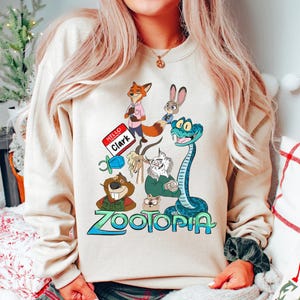 Puede incluir: Sudadera beige con un gr&aacute;fico colorido de personajes de la pel&iacute;cula animada "Zootopia". El dise&ntilde;o incluye un zorro, un conejo, una serpiente y otros animales, con el t&iacute;tulo de la pel&iacute;cula en azul y verde. Una etiqueta con el nombre dice "Hello Clark."