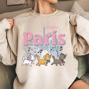 Puede incluir: Sudadera beige con la palabra "Par&iacute;s" en rosa, una ilustraci&oacute;n azul de la Torre Eiffel y tres gatos de dibujos animados. La palabra "Bonjour" est&aacute; encima de la palabra "Par&iacute;s". Los gatos son blancos, naranjas y grises.