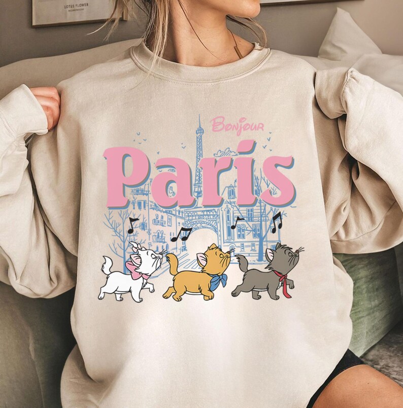 Puede incluir: Sudadera beige con la palabra "Par&iacute;s" en rosa, una ilustraci&oacute;n azul de la Torre Eiffel y tres gatos de dibujos animados. La palabra "Bonjour" est&aacute; encima de la palabra "Par&iacute;s". Los gatos son blancos, naranjas y grises.