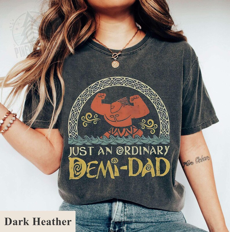 Op de afbeelding: Donkergrijs t-shirt met een vintage-stijl grafische afbeelding van een cartoonfiguur met spieren, een kroon en de tekst "Just an ordinary Demi-Dad".