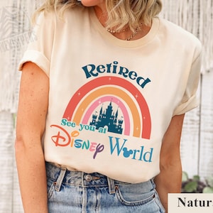 K&ouml;nnte beinhalten: Ein wei&szlig;es T-Shirt mit einem Regenbogen-Design und dem Text "Retired See you at Disney World" in blauer und schwarzer Schrift. Der Regenbogen hat eine Schloss-Silhouette in der Mitte. Das Shirt wird von einem Model getragen, das eine blaue Jeans tr&auml;gt.