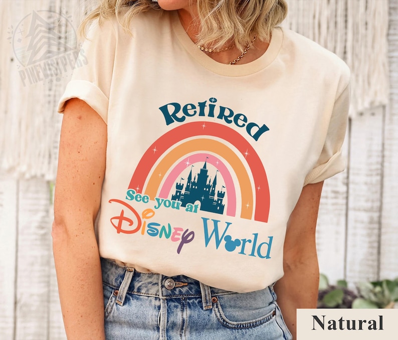 K&ouml;nnte beinhalten: Ein wei&szlig;es T-Shirt mit einem Regenbogen-Design und dem Text "Retired See you at Disney World" in blauer und schwarzer Schrift. Der Regenbogen hat eine Schloss-Silhouette in der Mitte. Das Shirt wird von einem Model getragen, das eine blaue Jeans tr&auml;gt.