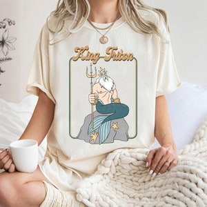 May include: A cream coloured t-shirt with a retro-style graphic of King Triton from The Little Mermaid. The graphic features King Triton sitting on a rock with his trident and a blue tail.