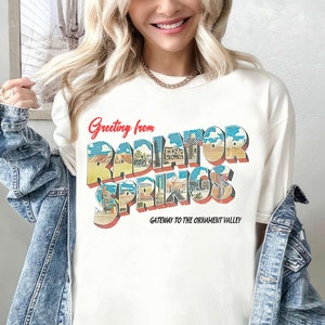 K&ouml;nnte beinhalten: Wei&szlig;es T-Shirt mit einer Grafik im Stil einer Vintage-Postkarte mit der Aufschrift "Greeting from Radiator Springs, Gateway to the Ornament Valley".