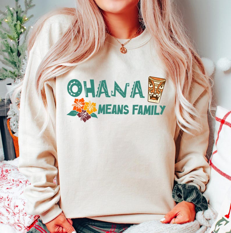 May include: Beige sweatshirt with the text "OHANA MEANS FAMILY" in teal and a tiki graphic. The design includes colorful hibiscus flowers. A cozy and casual top for family gatherings.