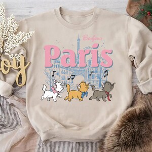 Puede incluir: Sudadera beige con un gr&aacute;fico de tres gatos de dibujos animados frente a una escena parisina con la Torre Eiffel. El texto "Bonjour Paris" est&aacute; encima de los gatos en rosa y azul.