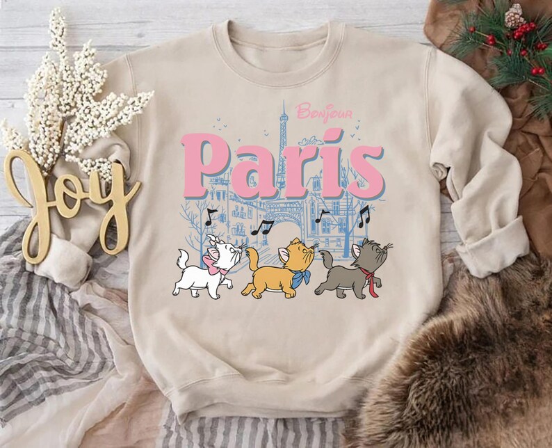 Puede incluir: Sudadera beige con un gr&aacute;fico de tres gatos de dibujos animados frente a una escena parisina con la Torre Eiffel. El texto "Bonjour Paris" est&aacute; encima de los gatos en rosa y azul.