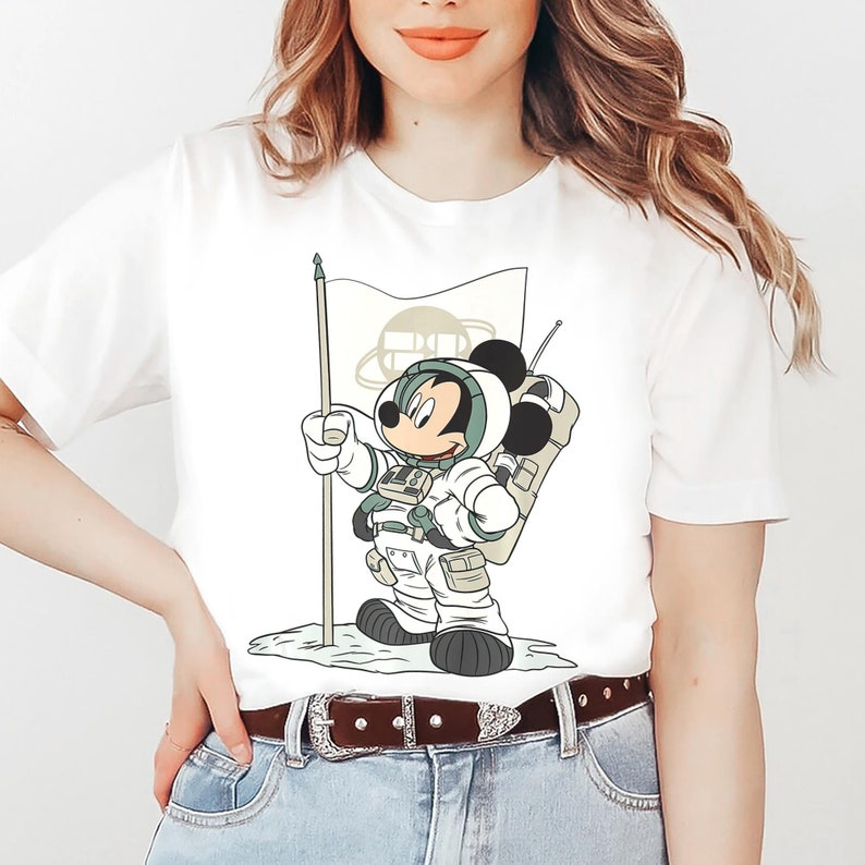 May include: White t-shirt with a cartoon image of Mickey Mouse dressed as an astronaut holding a flag with a planet on it. The astronaut is wearing a white spacesuit with green accents and a backpack.