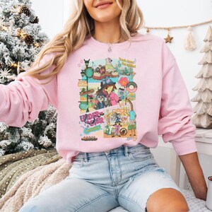 May include: A light pink sweatshirt featuring a colorful graphic with the text "Under Cover" and "Track down the spirit!". The graphic includes cartoon characters and various elements. The sweatshirt has long sleeves.