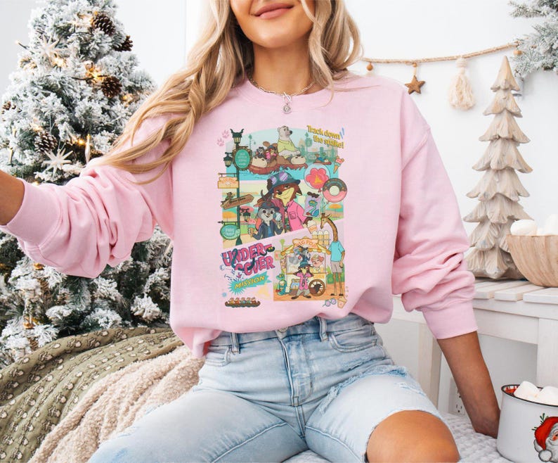 May include: A light pink sweatshirt featuring a colorful graphic with the text "Under Cover" and "Track down the spirit!". The graphic includes cartoon characters and various elements. The sweatshirt has long sleeves.