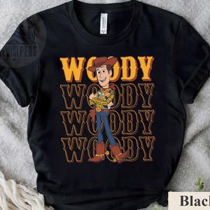 Puede incluir: Camiseta negra con una ilustraci&oacute;n de dibujos animados de Woody de Toy Story. El dise&ntilde;o incluye el nombre "WOODY" en amarillo y naranja repetido verticalmente. Woody est&aacute; representado con su atuendo de vaquero.