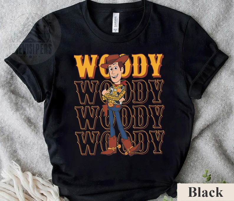 Puede incluir: Camiseta negra con una ilustraci&oacute;n de dibujos animados de Woody de Toy Story. El dise&ntilde;o incluye el nombre "WOODY" en amarillo y naranja repetido verticalmente. Woody est&aacute; representado con su atuendo de vaquero.