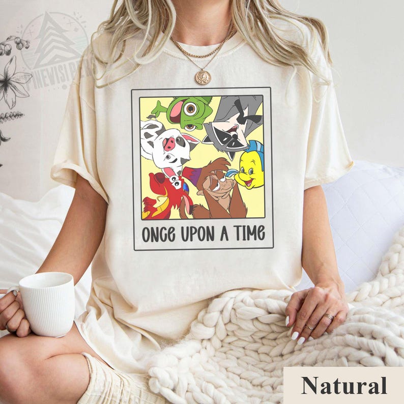 Op de afbeelding: Cr&egrave;mekleurig T-shirt met een ingelijste afbeelding van stripfiguren, waaronder een varken, een wasbeer en een vis, met de tekst "ONCE UPON A TIME" in het zwart. Het shirt is gemaakt van een zacht materiaal.