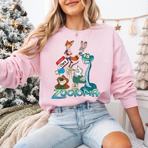 Puede incluir: Sudadera rosa claro con un dise&ntilde;o gr&aacute;fico colorido de personajes de la pel&iacute;cula Zootopia. El dise&ntilde;o incluye un zorro, un conejo, una serpiente y otros personajes animales. La palabra "Zootopia" est&aacute; impresa en azul.