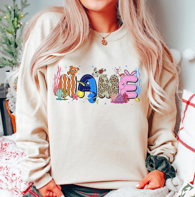 May include: Beige sweatshirt with the word "NAME" in colorful, cartoon-style lettering. The letters are designed with ocean-themed elements, including fish, coral, and a starfish. The sweatshirt has long sleeves.