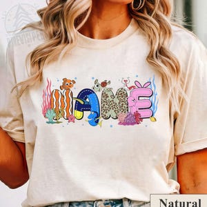 May include: A beige t-shirt featuring the word "NAME" in a colorful, aquatic-themed design. The letters are decorated with cartoon fish, coral, and other sea creatures. The word is in a playful, cartoonish font.