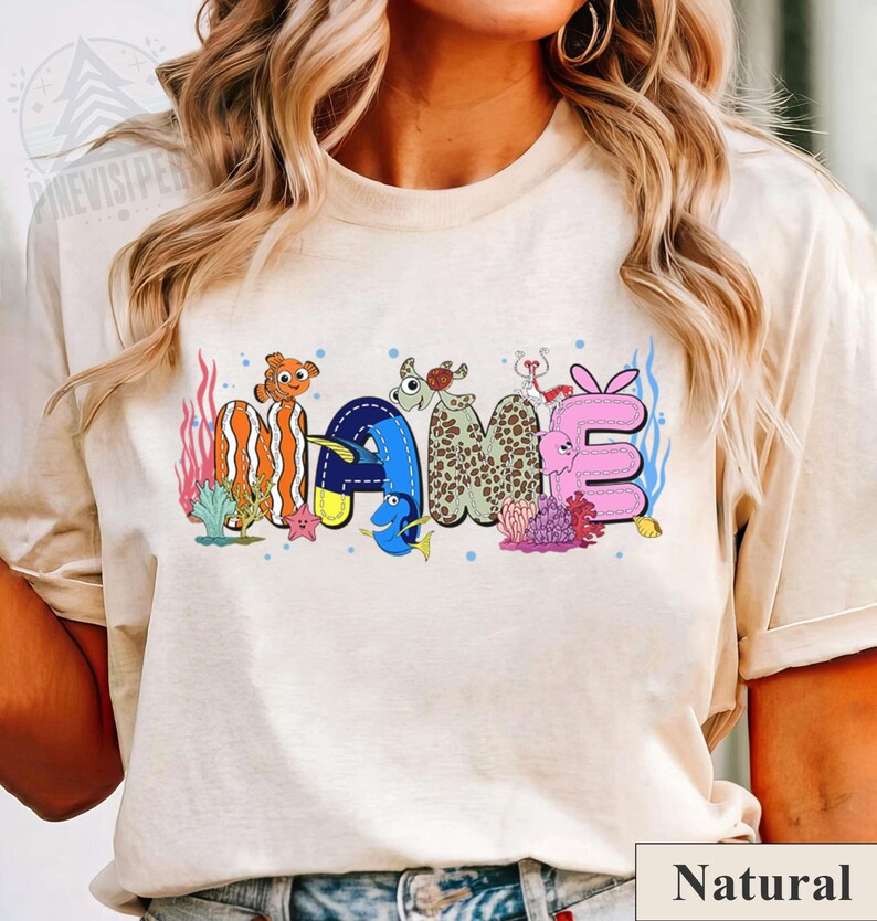 May include: A beige t-shirt featuring the word "NAME" in a colorful, aquatic-themed design. The letters are decorated with cartoon fish, coral, and other sea creatures. The word is in a playful, cartoonish font.